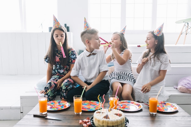 Foto gratuita il ragazzo e le ragazze che soffia i corni del partito si avvicinano alla torta