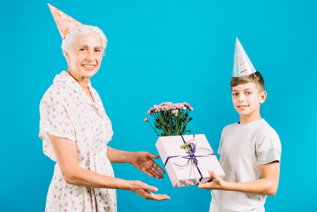 Free photo boy giving birthday gift and flowers to happy grandmother on blue backdrop