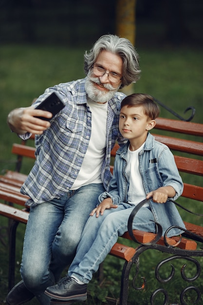 Мальчик и дедушка сидят на скамейке. Семья в парке. Старик играет с внуком. Дедушка пользуется телефоном.