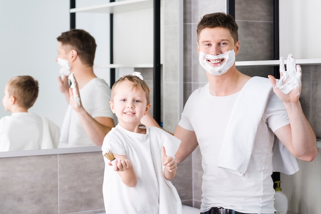 Boy holding brush in hand showing thumb up sign standing near his father with shaving foam on his face and hand