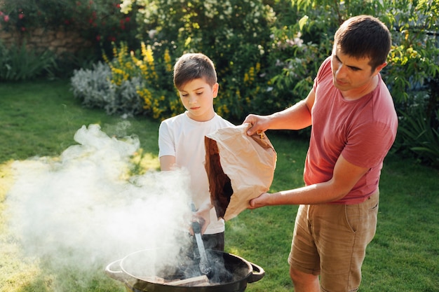 Мальчик держит щипцы стоя с его отцом, положить уголь в барбекю