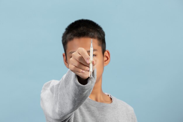 A boy is holdimg thermometer with gray long-sleeve shirt on blue wall.