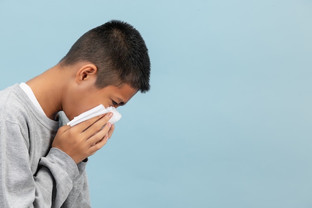A boy is sneezing into tissue and feeling sick on blue wall.