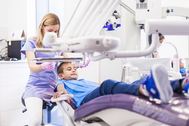 Мальчик, опираясь на стоматологический стул, получает лечение от женского стоматолога