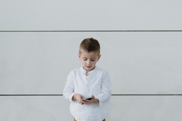 Boy looking down, playing on his phone with a white wall