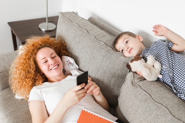 Free photo boy lying on sofa near her happy mother using cellphone