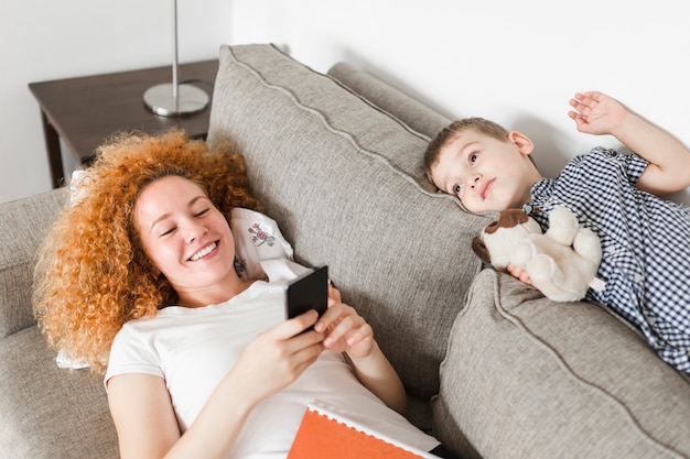 携帯電話を使って幸せな母親の近くのソファに横たわっている少年