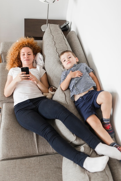 Boy lying on sofa with her mother using smartphone