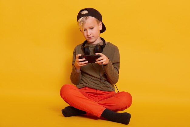 Boy playing game via cell phone, adorable male kid sitting isolated on yellow and holding mobile, guy dresses casually, posing with headphones around neck, keeping legs crossed.