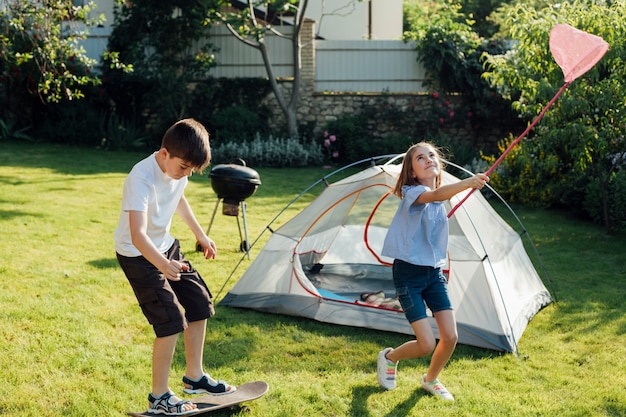 Boy playing skateboard near his sister catching butterflies and bugs with her scoop-net