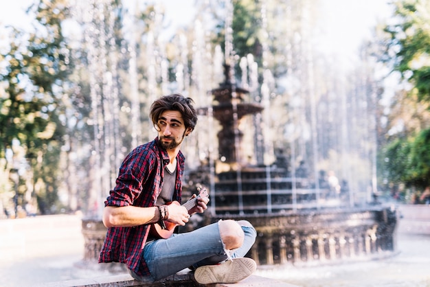 Free photo boy playing the ukelele sitting on a fountain