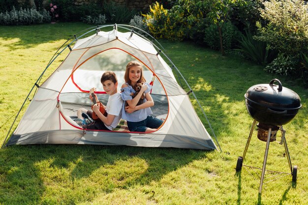 Boy playing ukulele sitting back to back his sister in tent near barbecue grill