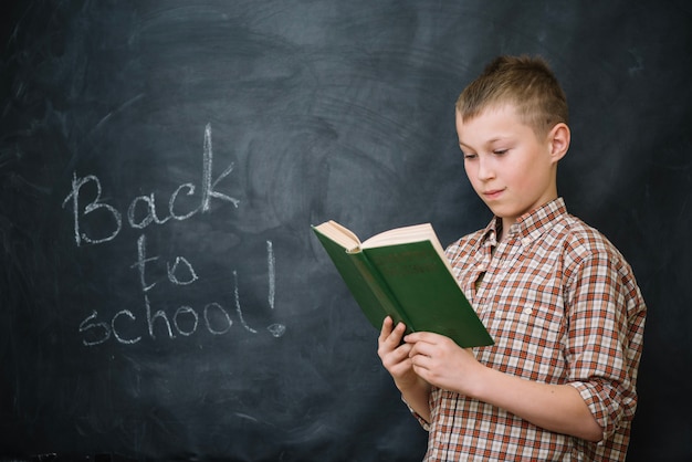 Boy reading book standing against blackboard