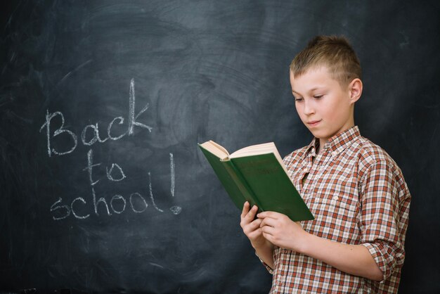 Boy reading book standing against blackboard