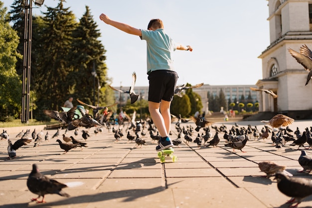 A boy riding penny board in the park on a warm summer time with pigeons and sky