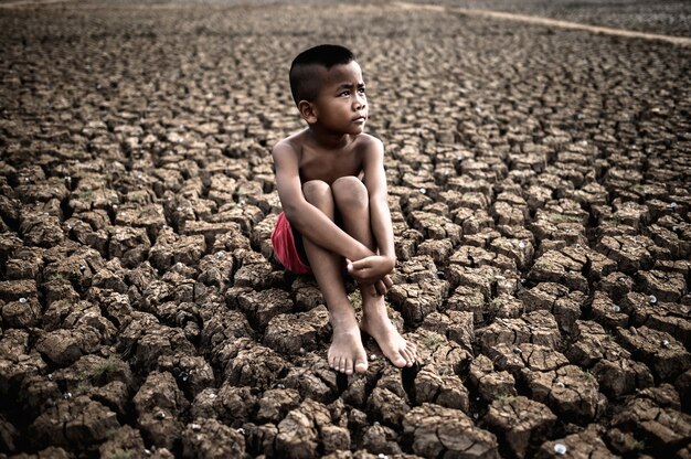 The boy sit hugging their knees bent and looking at the sky to ask for rain on dry soil.