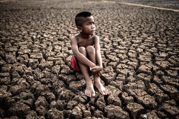 Мальчик сидит, обхватив колени согнутыми и глядя на небо, чтобы попросить дождя на сухой почве.