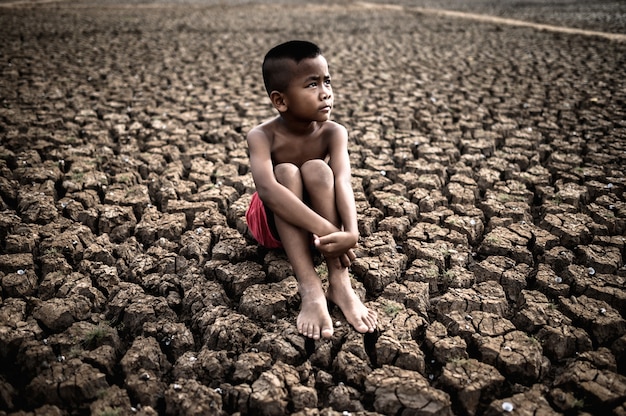 The boy sit hugging their knees bent and looking at the sky to ask for rain on dry soil.