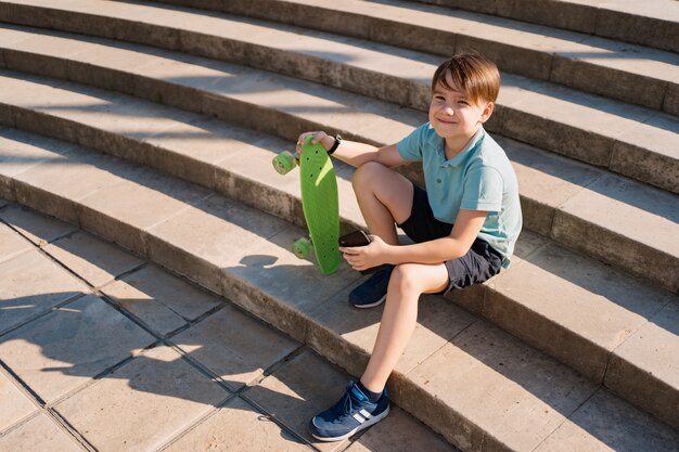 Boy sitting on stairs with smartphone in his hand and green penny board watching funny videos
