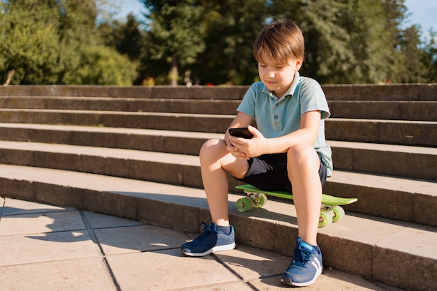 スマートフォンを手にして階段に座っている少年と面白い動画を見て緑のペニーボード