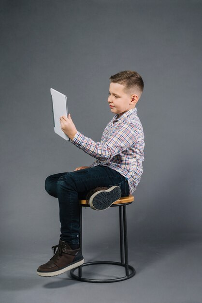 灰色の背景に対してデジタルタブレットを見てスツールに座っている男の子