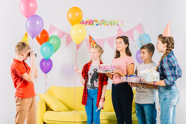 Мальчик фотографирует своих друзей с тортом на день рождения; подарок и воздушные шары