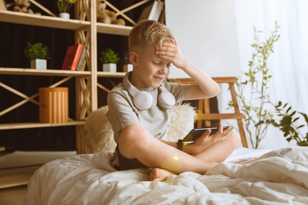 Boy using different gadgets at home. Little model with smart watches, smartphone or tablet and headphones. Making selfie, chating, gaming, watching videos. Interaction of kids and modern technologies.