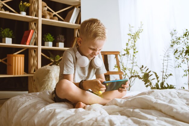 Мальчик дома с помощью различных гаджетов. Маленькая модель с умными часами, смартфоном или планшетом и наушниками.