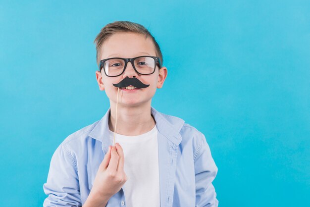 A boy wearing black eyeglasses holding black moustache prop in front of his upper lips