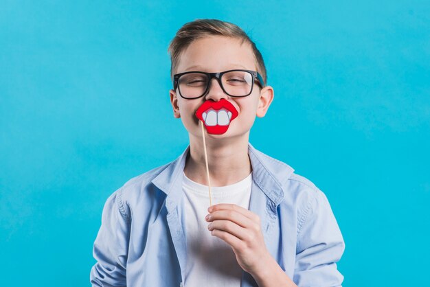 A boy wearing black eyeglasses holding smiling prop in front of his mouth against blue background