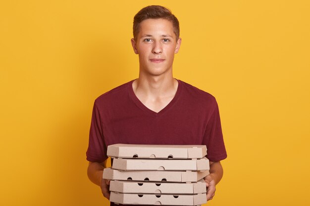 Мальчик нося maroon вскользь футболку поставляя коробки для пиццы, представляя изолированный на желтом цвете, смотря камеру, выглядит серьезной, молодой женщиной работая как работник доставляющий покупки на дом, делая его работу. Концепция людей