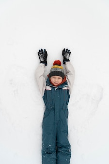 Boy in winter jumpsuit and knitted hat lies on the snow and makes snow angel. child walks outside in winter.
