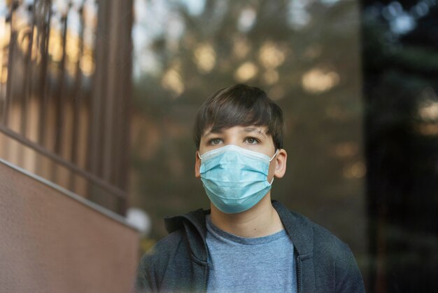 Boy with medical mask looking outside next to a window