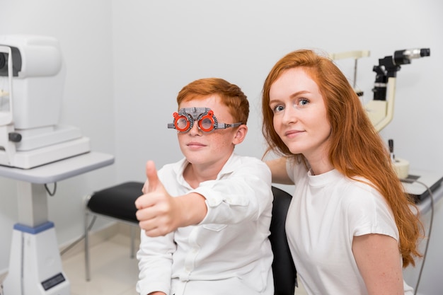 Boy with optometrist trial frame showing thumb up gesture sitting with young female ophthalmologist in clinic