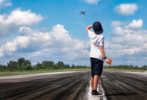 A boy with a paper airplane on the airstrip looking at the sky where a real plane is flying