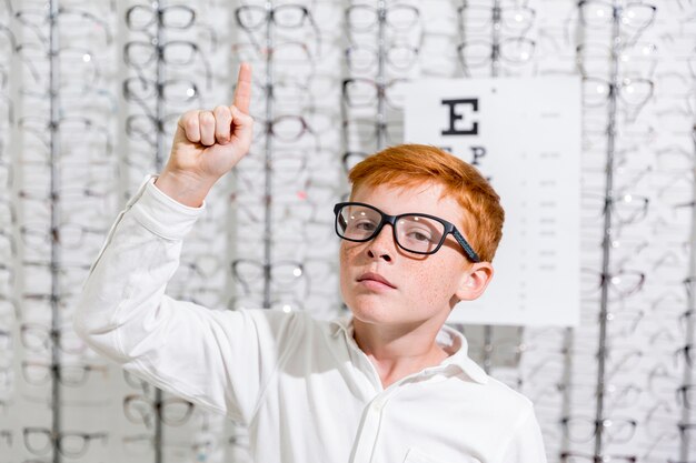 Boy with spectacle pointing at upward direction standing against eyeglasses display background