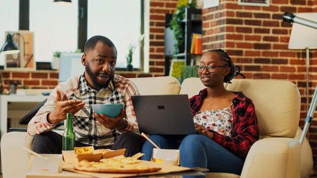 Бесплатное фото Парень и девушка смотрят сериал дома, едят чипсы и используют ноутбук для просмотра интернета. мужчина и женщина веселятся с шоу или фильмом, отдыхают на телевидении. съемка со штатива.