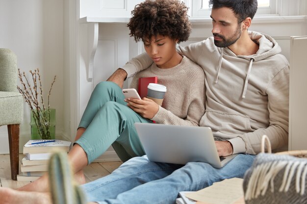 Boyfriend and girlfriend have rest after studying, view photos in social networks, use modern technologies for entertaiment sit on floor in modern apartment enjoy fresh drink from disposable paper cup
