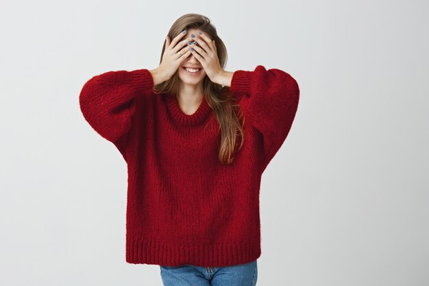 Boyfriend prepared some great present for his love. Studio shot of emotive charming woman in trendy clothes covering eyes with hands while smiling broadly, waiting to receive something good