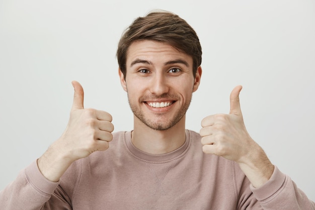 Foto gratuita il ragazzo supporta la fidanzata durante la partita. felice ragazzo europeo attraente con setole in abiti alla moda che sorride allegramente e mostra i pollici in su gesto