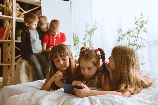 Boys and girls using different gadgets at home. Childs with smart watches, smartphone and headphones. Making selfie, chating, gaming, watching videos. Interaction of kids and modern technologies.
