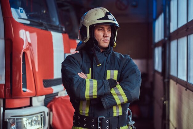 Храбрый пожарный в защитной форме стоит рядом с пожарной машиной в гараже пожарной части, скрестив руки и глядя в сторону
