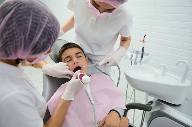 Brave school boy sitting on dentist's chair receiving medical treatment of his oral cavity by pediatrician dentist and his assistant in modern dentistry clinic