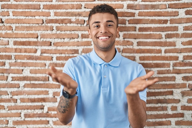 Brazilian young man standing over brick wall smiling cheerful offering hands giving assistance and acceptance.