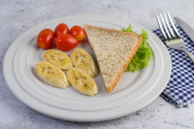 Bread, banana, and tomato on white plate with fork and a knife.