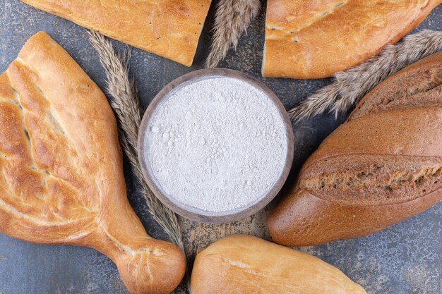 Bread loaves and wheat stalks around a bowl of flour on marble surface