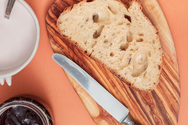 Bread slice and butter knife on chopping board with milk and jam on colored background