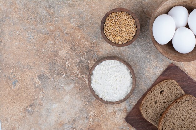 Bread slices, bowls of eggs, flour and barley on marble surface