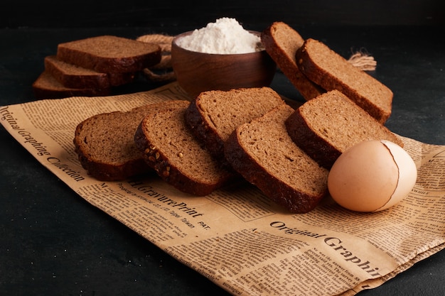 Bread slices,a cup of flour and an egg on the piece of newspaper. 