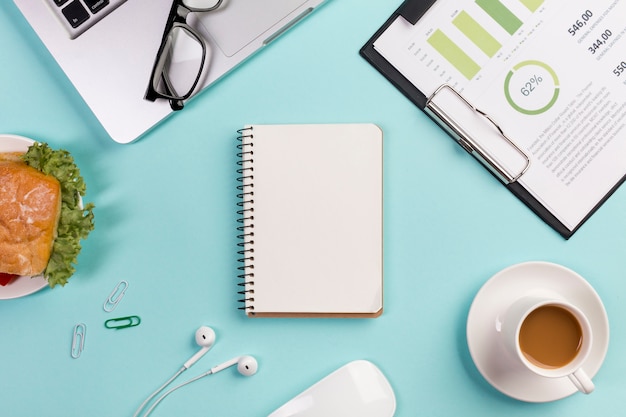 Breakfast,business chart,laptop,eyeglasses,spiral notepad,earphones and mouse on blue office desk